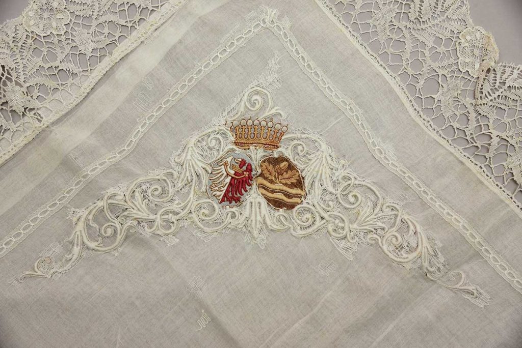 Polštář z jemného bílého etamínu zdobený paličkovanou krajkou a výšivkou s rodovými erby, vlevo Kolowratská orlice, vpravo erb rodu Khevenhüllerů, polovina 19. století
